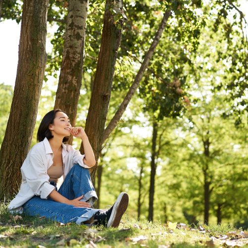 Relaxed young woman, resting near tree, sitting in park on lawn under shade, smiling and looking